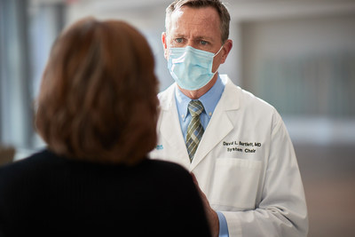 青いフェイスマスクを着け、青いシャツとストライプのネクタイの上に実験用白衣を着た男性が、黒いセーターを着た茶色い髪の女性に面して立っている