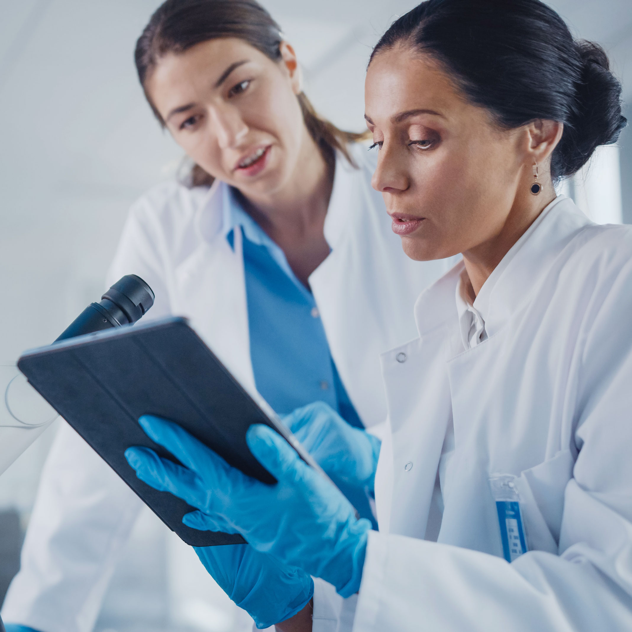 two women wearing white lab coats and blue gloves looking at a tablet