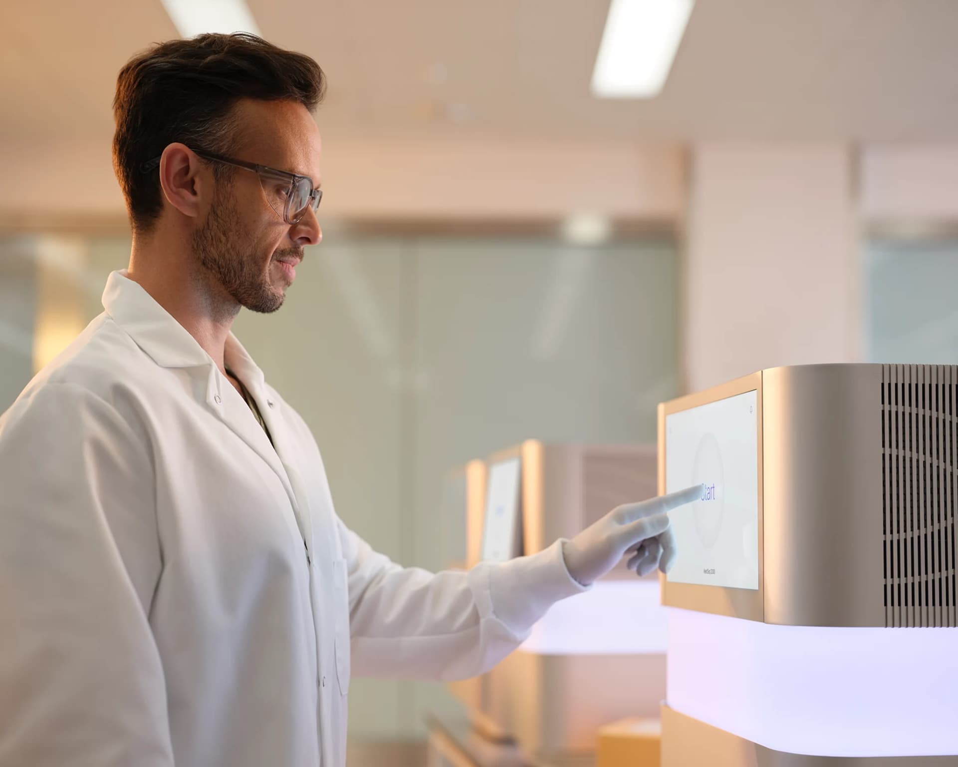Profile image of a male scientist selecting the start screen on a NextSeq 1000/2000 in a dry lab on a benchtop, other instruments and packaging blurry in the background.