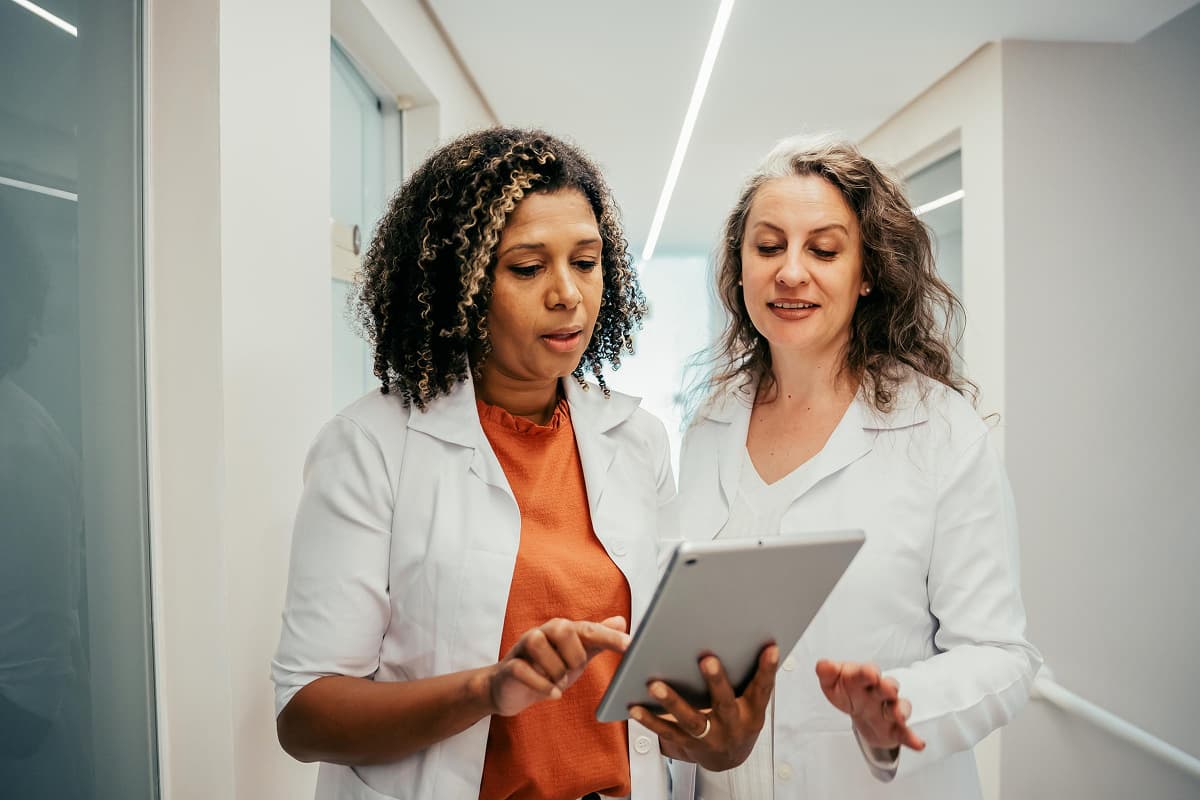 Two diverse female healthcare providers standing in an office hallway, one holding a tablet, having a discussion.