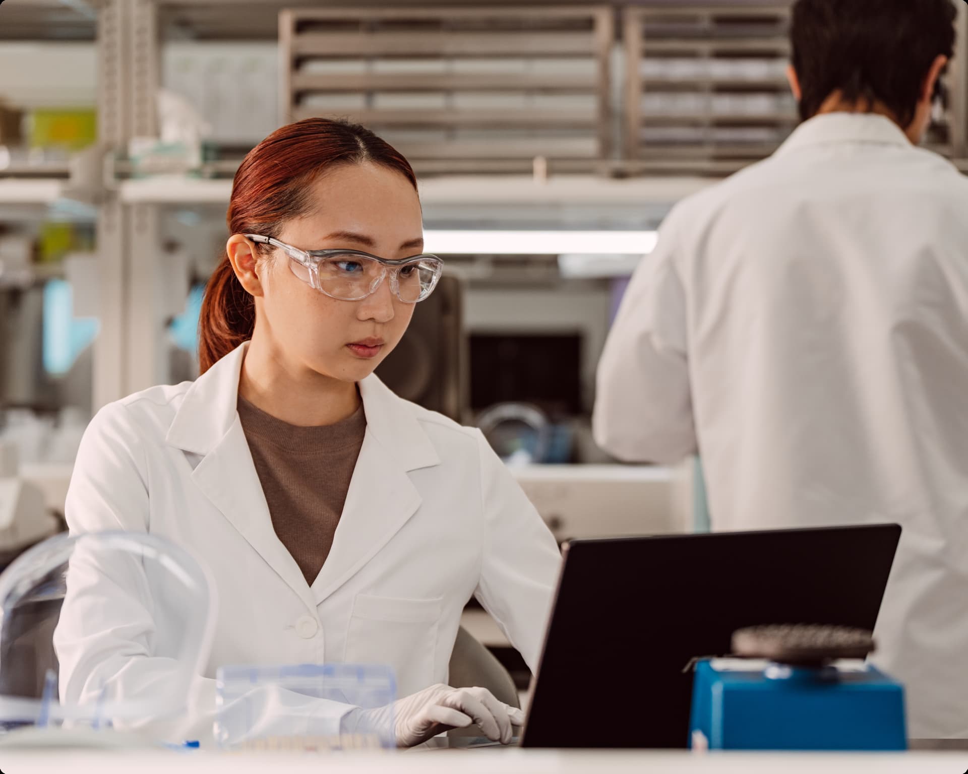 Female scientist interacting with a laptop in a lab setting; blurry image of a scientist in the background.