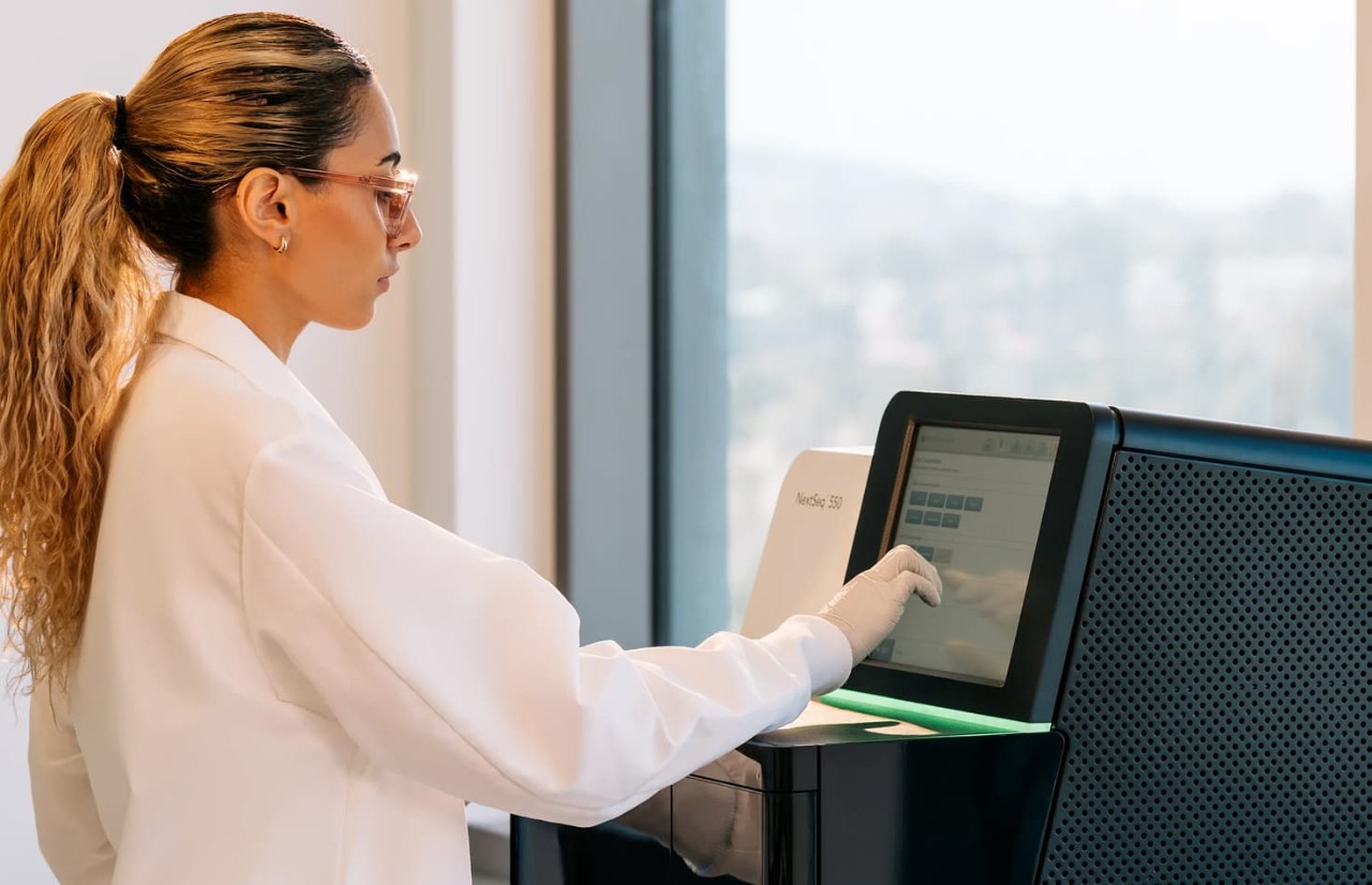 Profile view of a female scientist interacting with the load consumables touch screen on the monitor of a NextSeq 550 in front of a window; green status bar