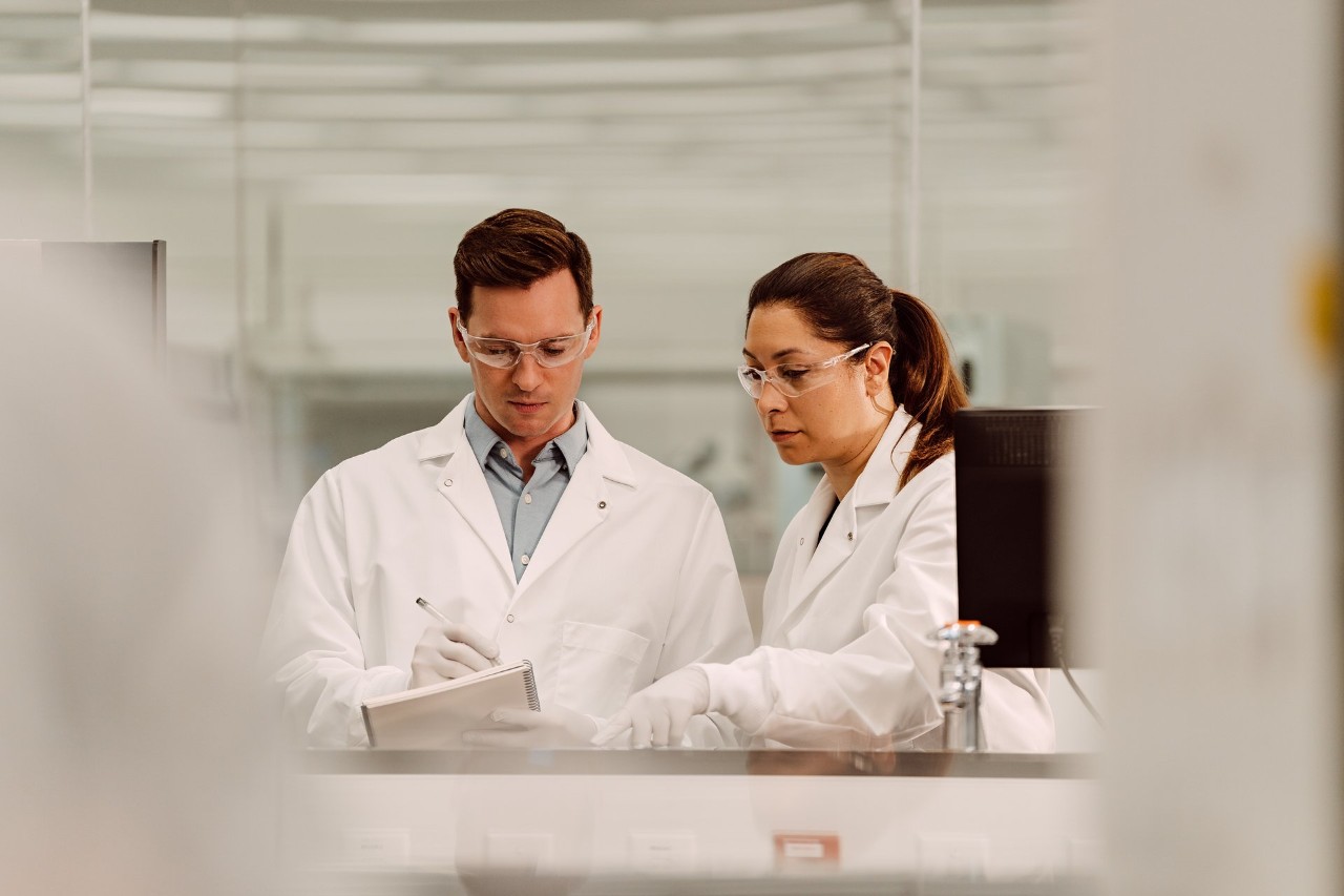 Two scientists, male and female, using a pen to write on a notepad while analyzing on a monitor, not visible, in a lab setting.