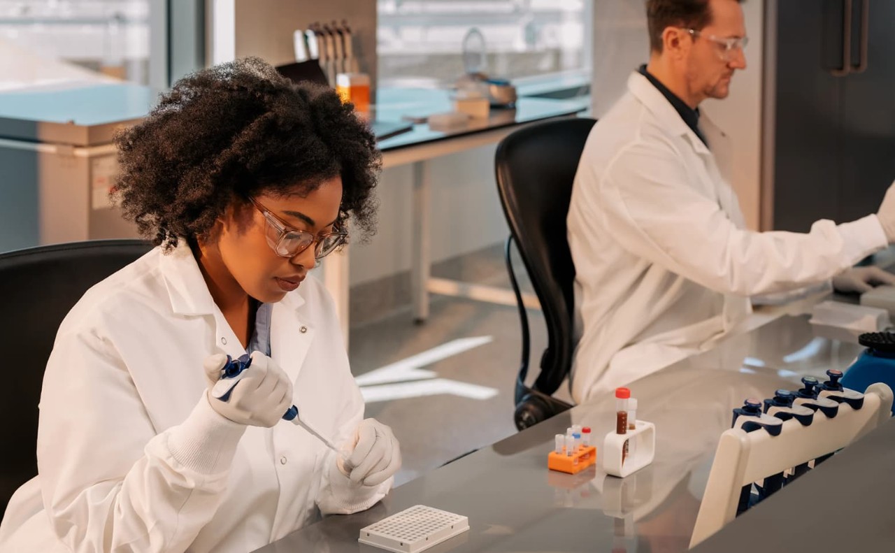 Side view of a female scientist using a single pipette; pipetting clear liquid in a sample tube; male scientist sitting next to the female working separately on the lab bench; also on the lab bench are six library tubes filled with clear liquid and and two larger tubes filled dark liquid; library prep box blurry in the background