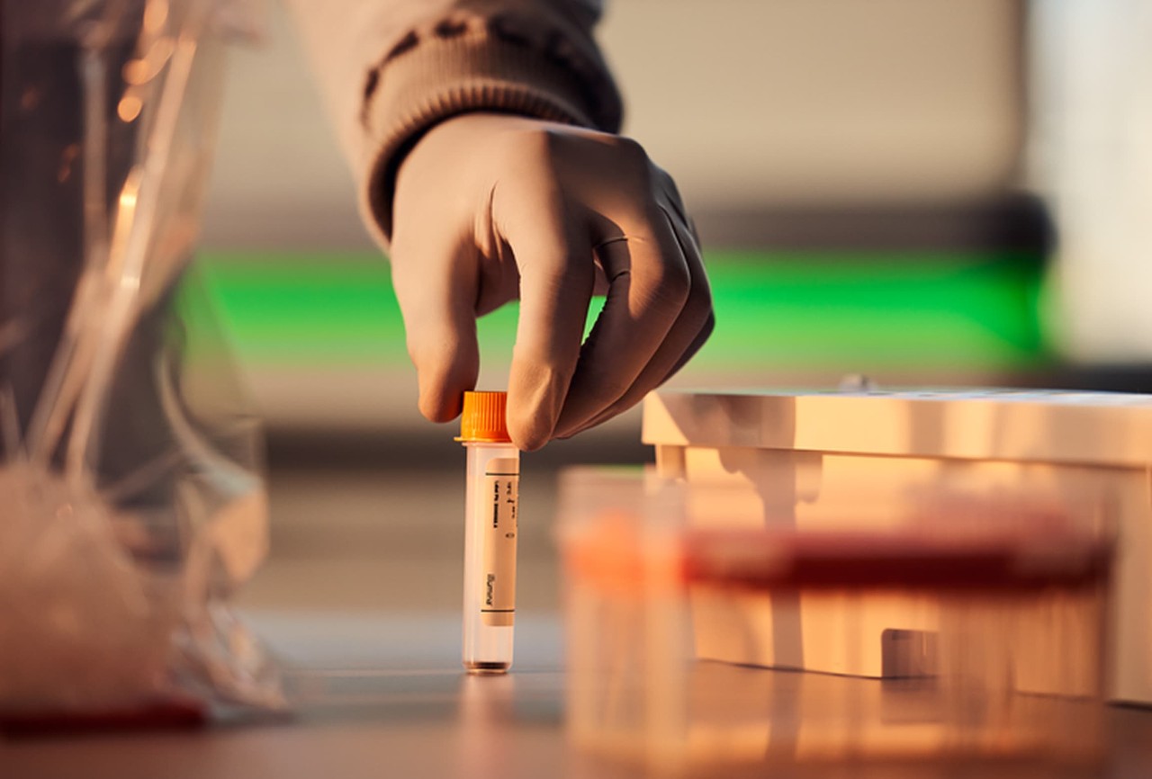 Scientist holding reagent tube with dark liquid, NovaSeq 6000 blurry in background