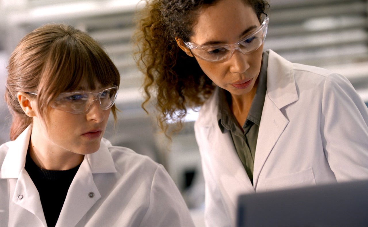 Two female scientists are working together, looking down at a monitor on a laptop in a lab setting.