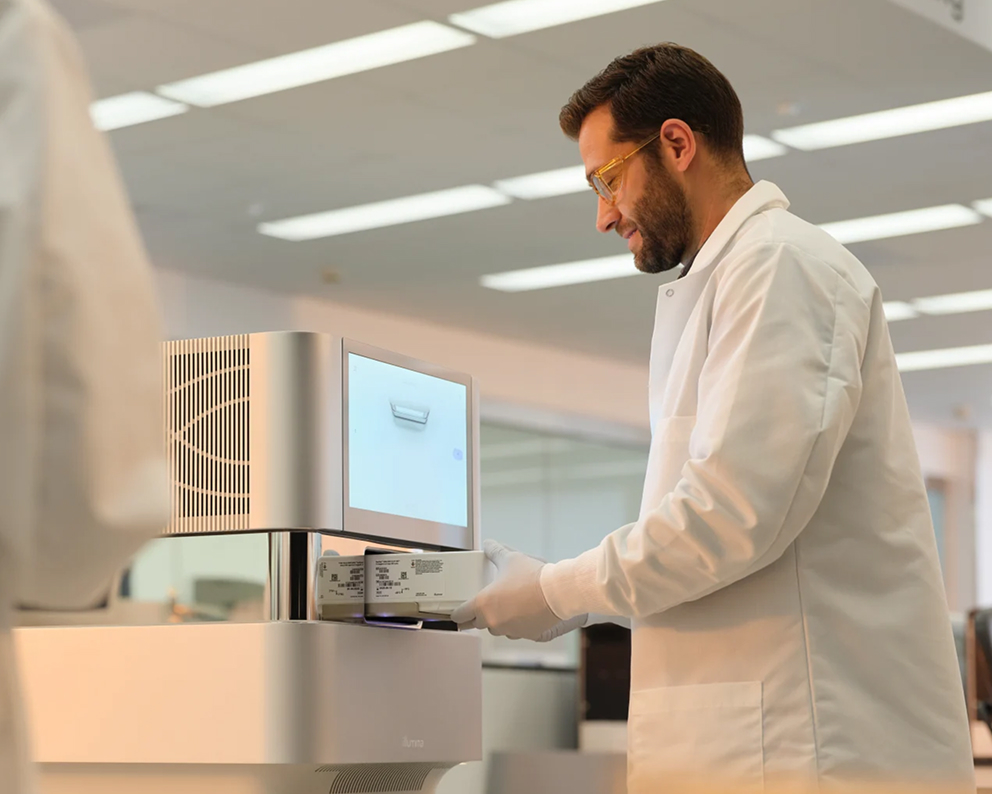 Profile view of male scientist standing in front of NextSeq 1000/2000 displaying load cartridge screen and inserting reagent cartridge. Blurry female scientist in the foreground.